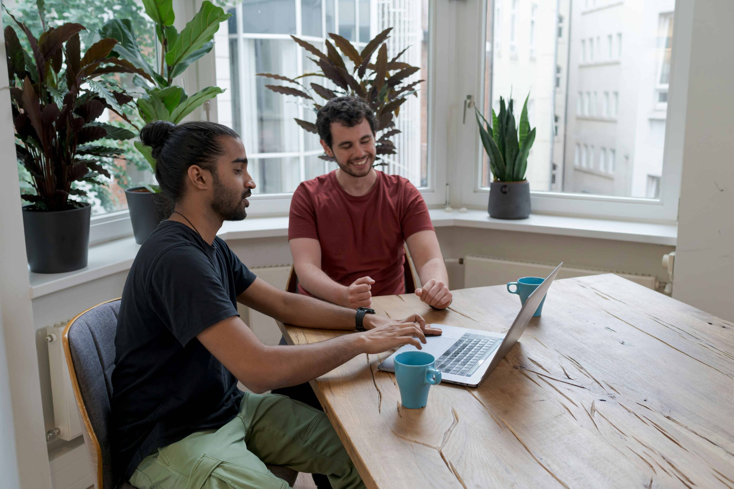 Twee mannen die samen achter een laptop aan een houten tafel werken, de een typend en de ander lachend.